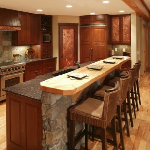 Island comprised of stone wall and rich wood paneling matching the cabinetry throughout this kitchen, features marble countertop and raised wood dining surface.